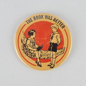 May include: A round, yellow button with a red circle in the center. The circle has the text "THE BOOK WAS BETTER" around it. Two children are depicted in black and white, one sitting in a hammock and reading a book, the other standing and looking at the reader.
