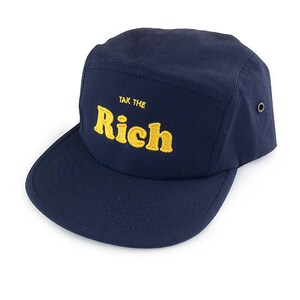 May include: A navy blue five-panel cap with a flat brim. The cap features the embroidered text "TAX THE RICH" in yellow. The cap has a structured design and a small metal eyelet on the side.