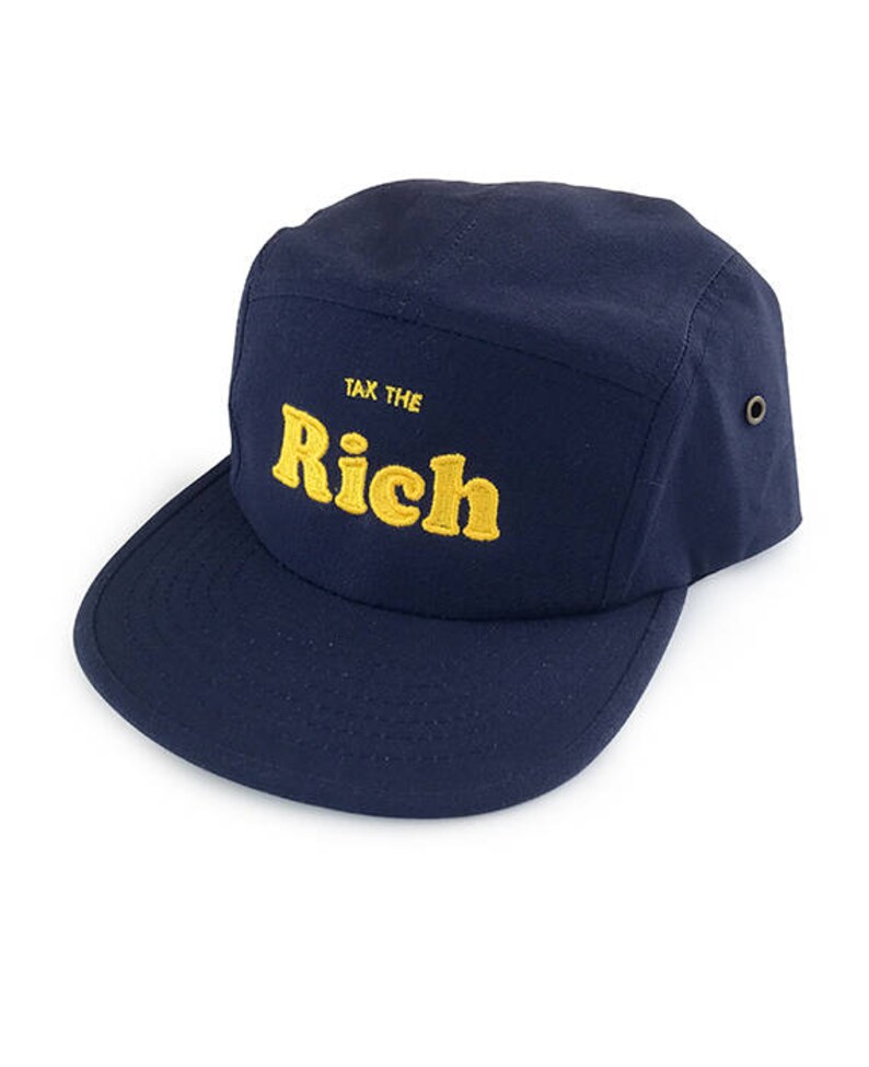 May include: A navy blue five-panel cap with a flat brim. The cap features the embroidered text "TAX THE RICH" in yellow. The cap has a structured design and a small metal eyelet on the side.