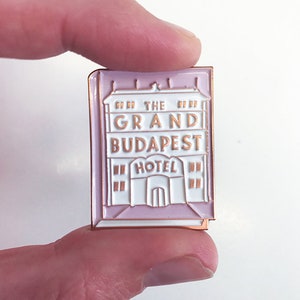 May include: A pink and white enamel pin featuring the Grand Budapest Hotel logo. The pin is shaped like a building with the words "The Grand Budapest Hotel" and "Hotel" written on it.