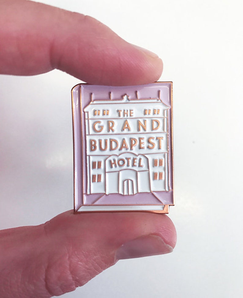 May include: A pink and white enamel pin featuring the Grand Budapest Hotel logo. The pin is shaped like a building with the words "The Grand Budapest Hotel" and "Hotel" written on it.