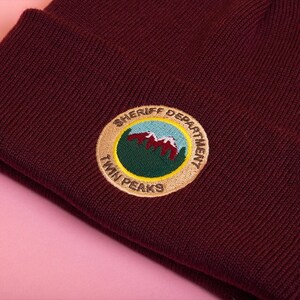 May include: A maroon beanie with an embroidered patch that says "Sheriff Department Twin Peaks" with a green mountain range and red peaks inside the circle.