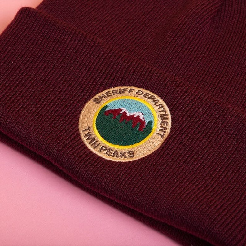 May include: A maroon beanie with an embroidered patch that says "Sheriff Department Twin Peaks" with a green mountain range and red peaks inside the circle.