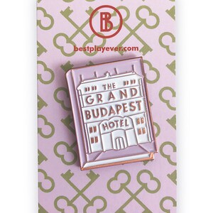 May include: A pink enamel pin featuring a white building with the text "The Grand Budapest Hotel" in gold lettering. The pin is on a pink background with a pattern of gold keys.