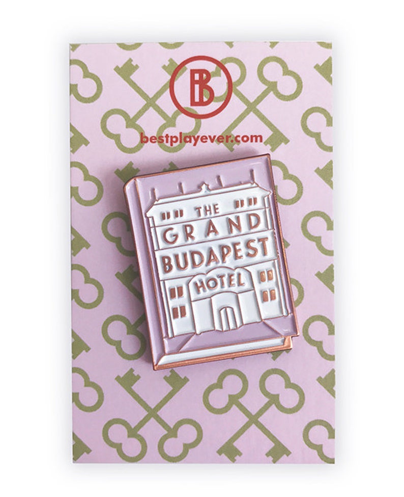 May include: A pink enamel pin featuring a white building with the text "The Grand Budapest Hotel" in gold lettering. The pin is on a pink background with a pattern of gold keys.