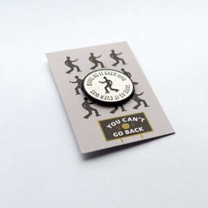 May include: A white enamel pin with a black outline featuring a silhouette of a person dancing with the text "Same as it ever was" written around the edge. The pin is displayed on a card with a black and white illustration of people dancing and the text "You can't go back".
