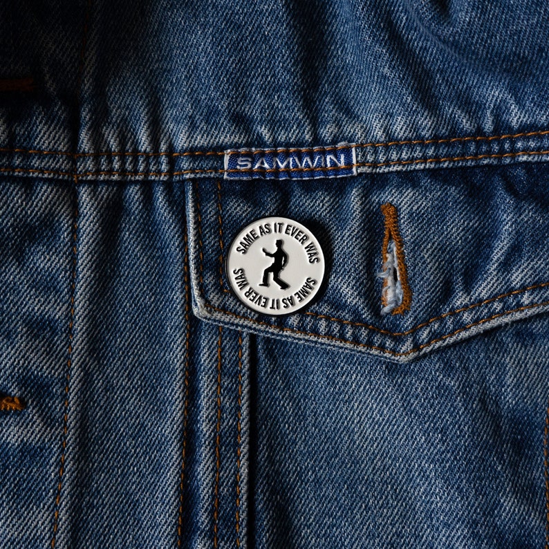 May include: A silver pin with the text "Same as it ever was" and a silhouette of a person dancing. The pin is on a blue denim jacket.