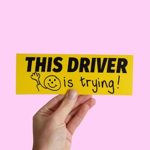 Puede incluir: Pegatina amarilla para parachoques con texto negro que dice "Este conductor lo está intentando!" y una cara sonriente de dibujos animados con la mano levantada.