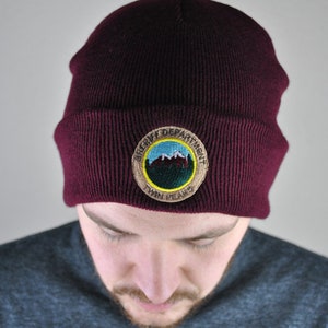 May include: A maroon beanie with a brown embroidered patch that says "Sheriff Department Twin Peaks". The patch features a blue mountain scene with white snow peaks.