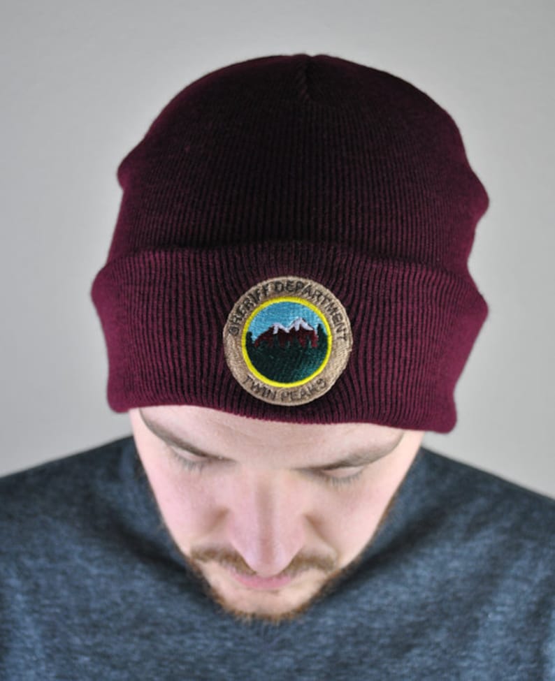 May include: A maroon beanie with a brown embroidered patch that says "Sheriff Department Twin Peaks". The patch features a blue mountain scene with white snow peaks.