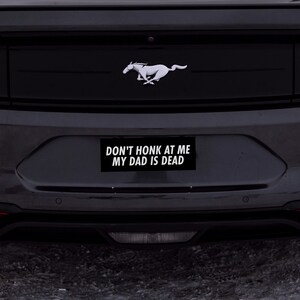 May include: A black car with a silver running horse emblem on the back. The car has a bumper sticker that reads "Don't honk at me my dad is dead."