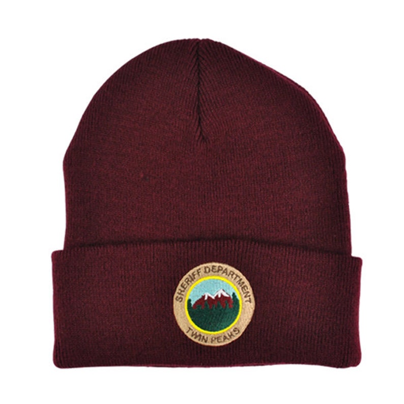 May include: A maroon beanie with a circular embroidered patch that says "Sheriff Department Twin Peaks". The patch features a mountain range and green trees.
