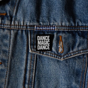 May include: A black and white enamel pin with the text "Dance Magic Dance" on a denim jacket.