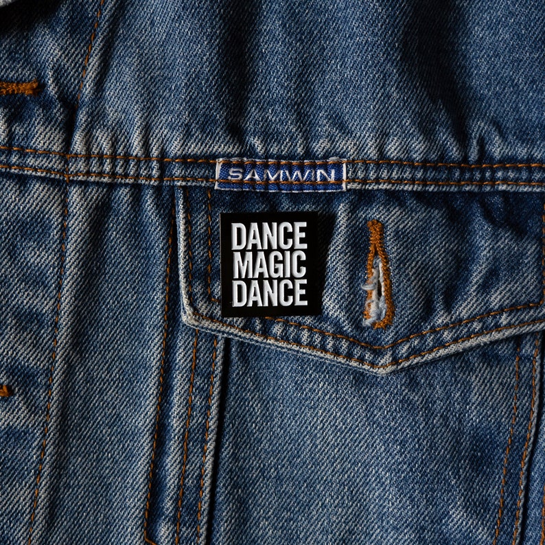 May include: A black and white enamel pin with the text "Dance Magic Dance" on a denim jacket.