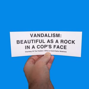 May include: A white rectangular sign with black text, held against a blue background. The text reads "VANDALISM: BEAUTIFUL AS A ROCK IN A COP'S FACE" with a smaller line below stating "Courtesy Of The Feederz: Office of Anti-Public Relations."