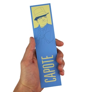 May include: A blue bookmark with a yellow illustration of Truman Capote wearing a hat and a yellow inscription that reads "CAPOTE".