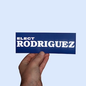Può includere: Un cartello rettangolare blu con testo bianco che dice "ELECT RODRIGUEZ".