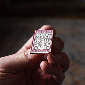 May include: A pink and gold enamel pin featuring a building with the text "The Grand Budapest Hotel".