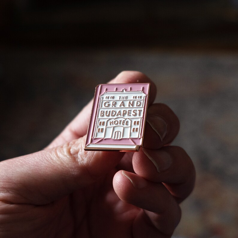 May include: A pink and gold enamel pin featuring a building with the text "The Grand Budapest Hotel".