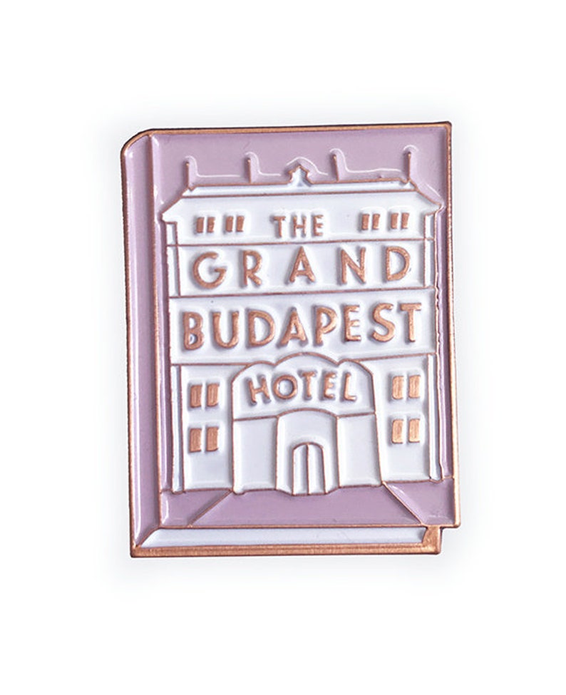 May include: A pink enamel pin featuring a white building with the text "The Grand Budapest Hotel" in gold lettering.