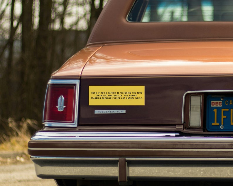 May include: A brown car with a yellow bumper sticker that reads "HONK IF YOU'D RATHER BE WATCHING THE 1999 CINEMATIC MASTERPIECE THE MUMMY STARRING BRENDAN FRASER AND RACHEL WEISZ".