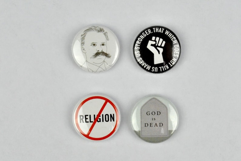 May include: Four round button pins with different designs. The first pin features a black and white illustration of a man with a mustache. The second pin has a black background with a white fist and the text "That which does not kill us makes us stronger." The third pin has a red circle with a diagonal line through it and the word "RELIGION" in black text. The fourth pin has a gray background with a white tombstone and the text "God is Dead."