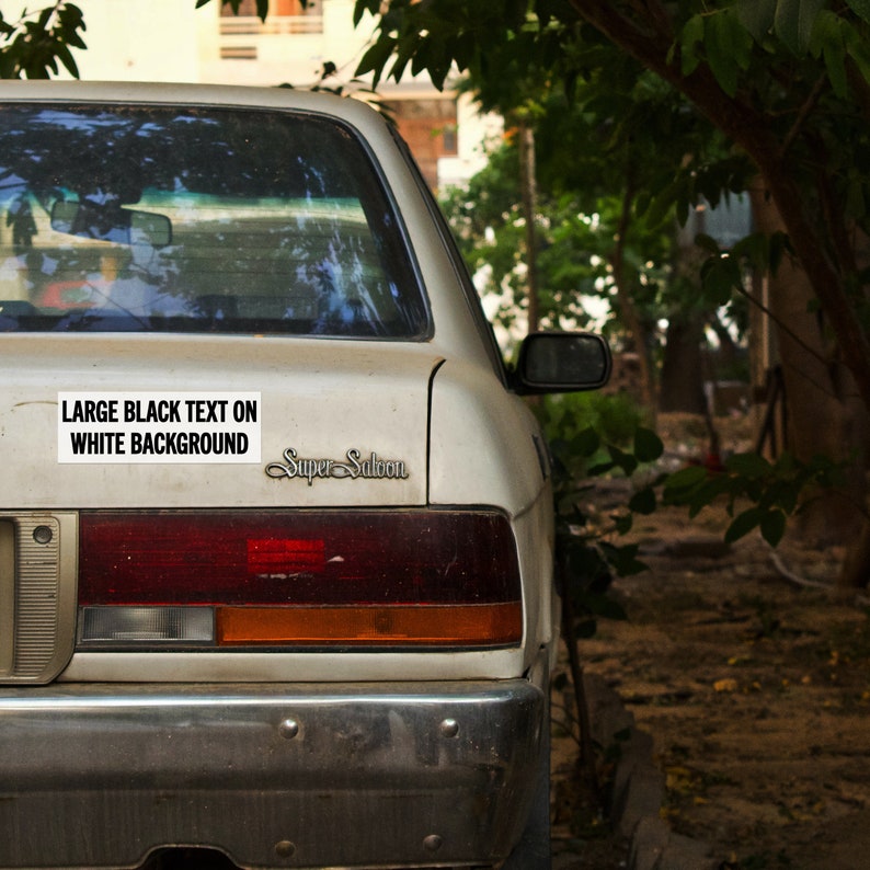 May include: A white car with a black bumper and a black sticker that says "LARGE BLACK TEXT ON WHITE BACKGROUND" on the back. The car also has a chrome "Super Saloon" emblem on the back.