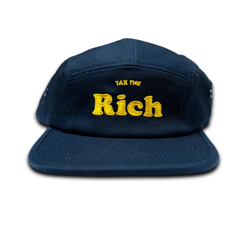 May include: A navy blue five-panel cap with a flat brim. The cap features the embroidered text "TAX THE RICH" in yellow thread. The hat is a casual accessory.