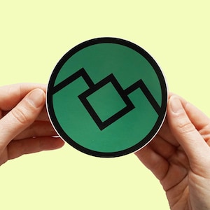 May include: A green and black circular sticker with a geometric design. The design features a black square inside a larger black square, with two black lines extending from the top and bottom of the larger square.