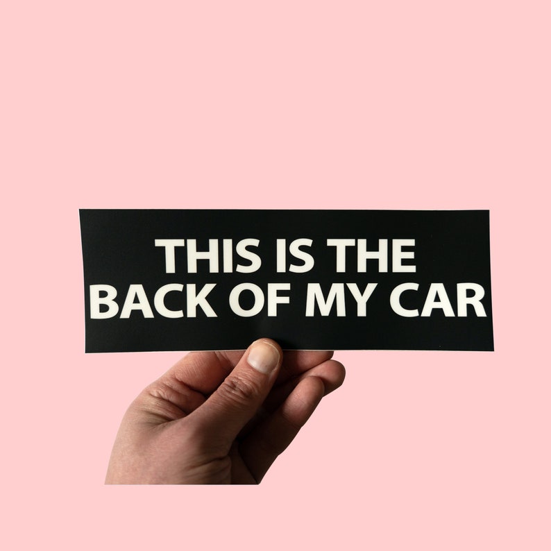 May include: Black rectangular bumper sticker with the text "THIS IS THE BACK OF MY CAR" in white, held against a light pink background. The sticker is held by a hand.