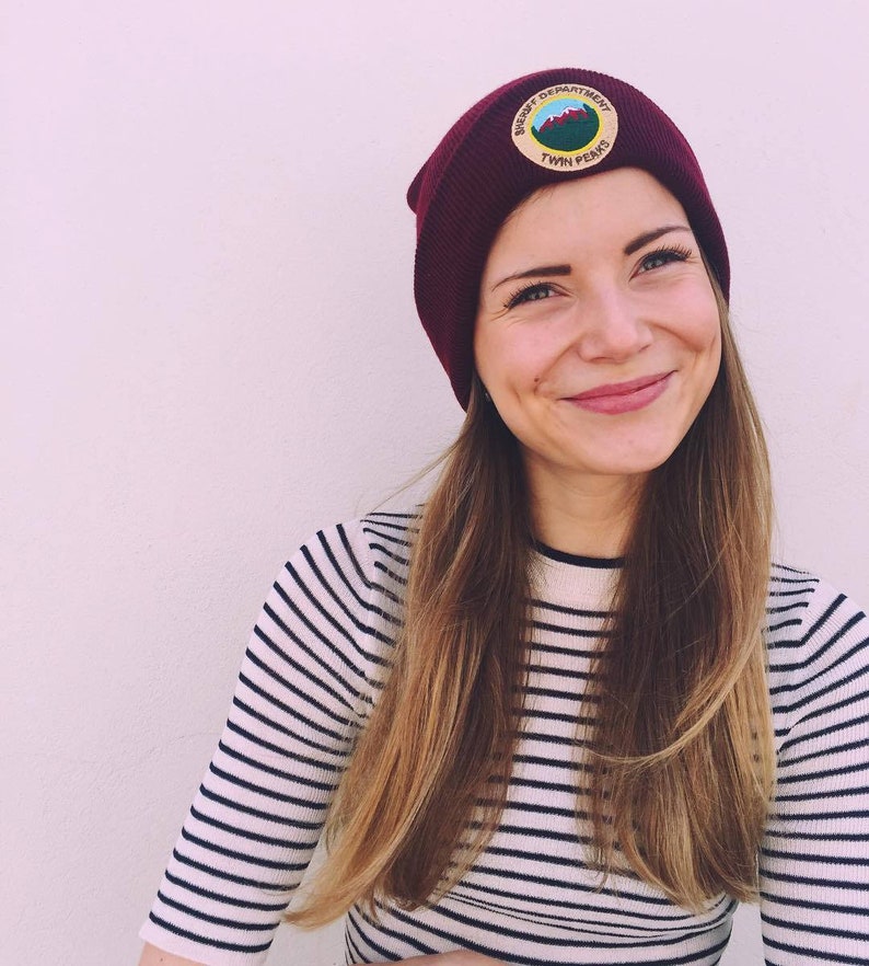 May include: A woman wearing a burgundy beanie with a patch that says "Sheriff Department Twin Peaks". She is wearing a white and black striped shirt.