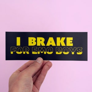 May include: A black and yellow bumper sticker with the text "I BRAKE FOR EMO BOYS" in a pixelated font.