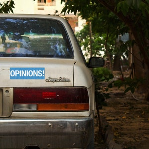 May include: A white car with a blue bumper sticker that says "OPINIONS!" and a chrome Super Saloon emblem. The car is parked in front of a tree.