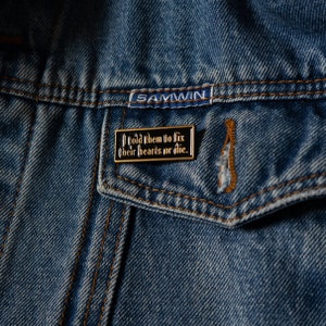 May include: A black and gold enamel pin with the text "I told them to fix their hearts or die." The pin is attached to a blue denim jacket.
