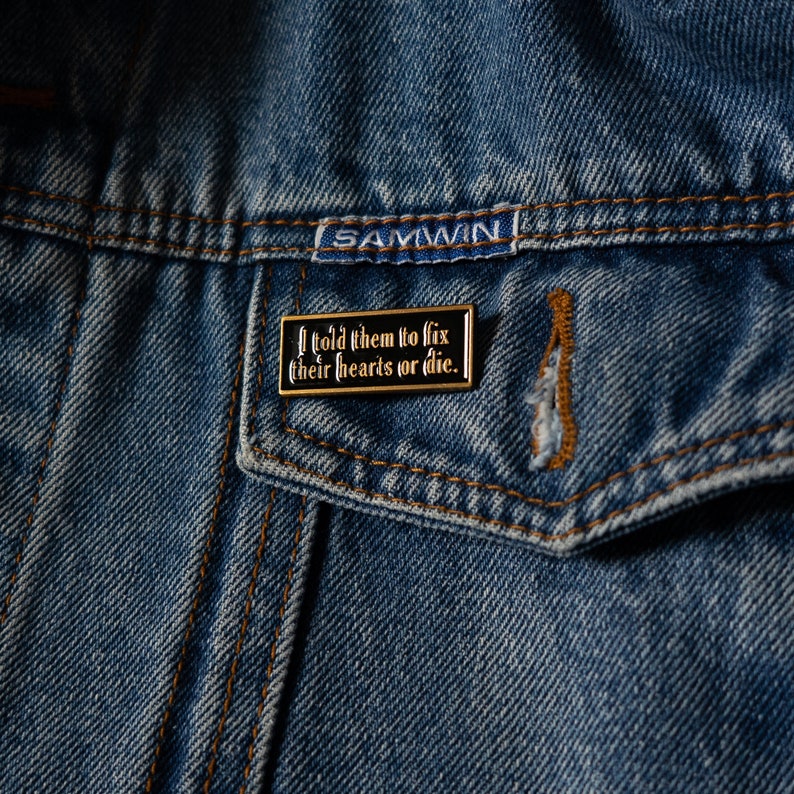 May include: A black and gold enamel pin with the text "I told them to fix their hearts or die." The pin is attached to a blue denim jacket.