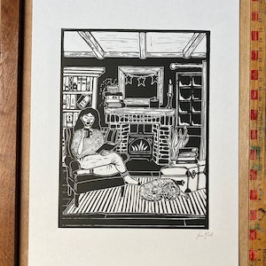 May include: Black and white linocut print of a woman sitting in a chair by a fireplace, reading a book. The woman is wearing a patterned sweater and has her hair pulled back. The fireplace has a fire burning in it and there is a cat curled up on the floor nearby. The print is signed "Jane Bird" in the bottom right corner.