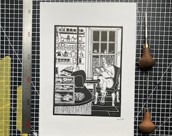 May include: Black and white linocut print of a woman sitting in a cafe, reading a book. The cafe has a counter with shelves of mugs and a window with a view of a street. The print is signed by the artist.