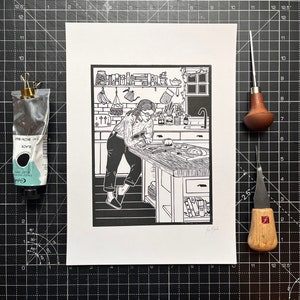 Puede incluir: Ilustración en blanco y negro de una persona en una cocina haciendo una pizza. La persona lleva una camisa de manga larga y pantalones. La cocina tiene una ventana, una encimera y un armario. Hay varios artículos de cocina en la encimera, incluyendo un tazón, un rodillo y un cortador de pizza.
