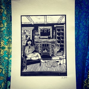 May include: A black and white linocut print of a woman sitting in a chair by a fireplace, reading a book. The fireplace has a fire burning in it and there are books stacked on a shelf to the left of the fireplace. The woman is wearing a long-sleeved shirt and has her hair pulled back. There is a dog sleeping on the floor in front of the fireplace.