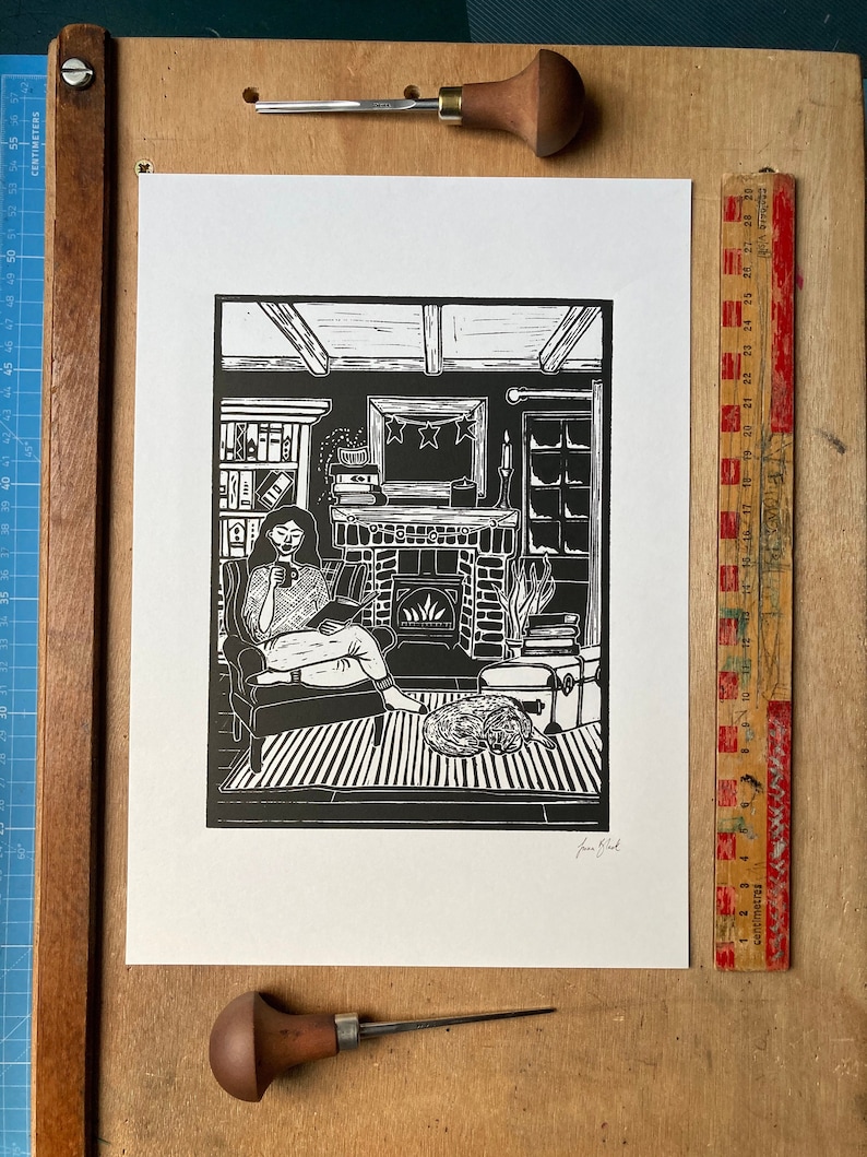 May include: Black and white linocut print of a woman sitting in a chair by a fireplace, reading a book. The woman is wearing a patterned sweater and has her hair pulled back. The fireplace has a fire burning in it and there is a cat curled up on the floor nearby. The print is signed "Jane Bird" in the bottom right corner.