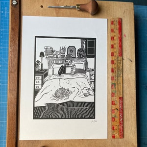 Peut inclure: Gravure sur linol&eacute;um en noir et blanc d'une personne lisant au lit avec un chat. L'impression est encadr&eacute;e et accroch&eacute;e &agrave; un mur.