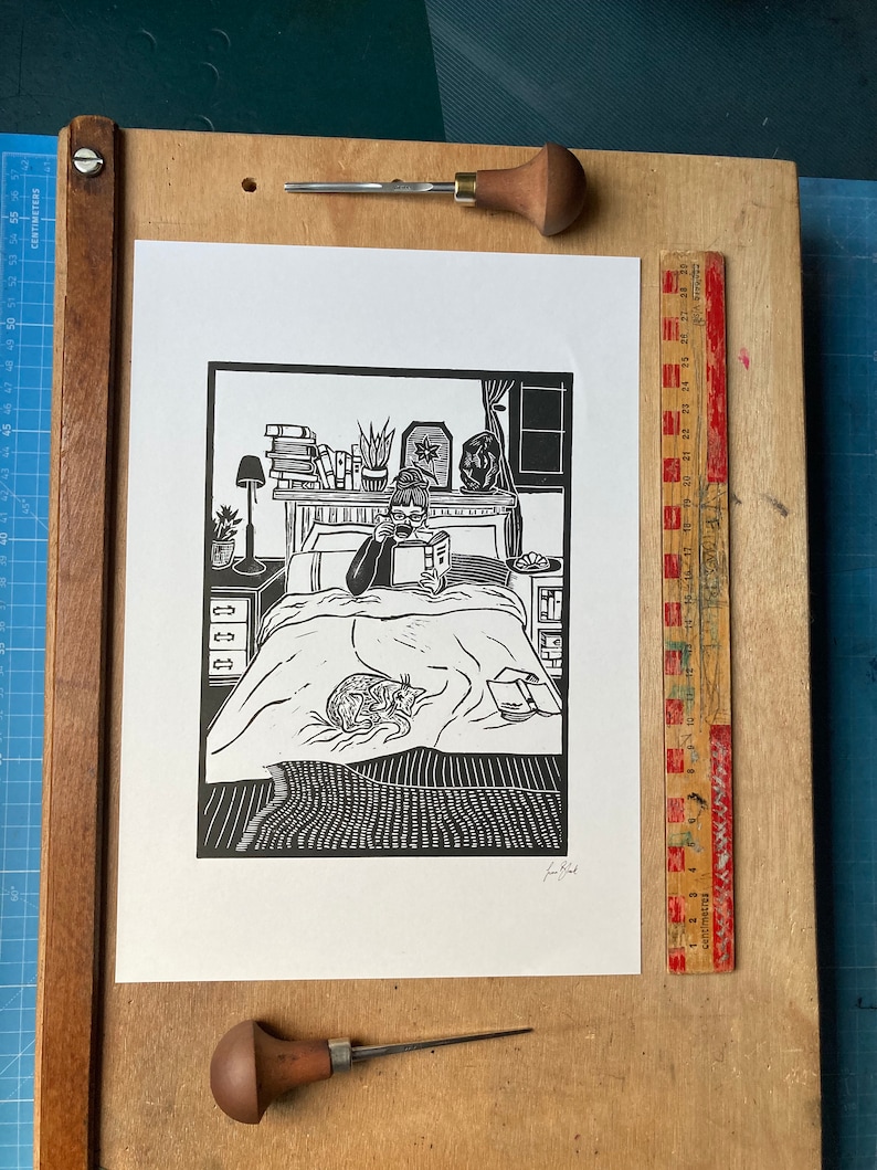 Peut inclure: Gravure sur linol&eacute;um en noir et blanc d'une personne lisant au lit avec un chat. L'impression est encadr&eacute;e et accroch&eacute;e &agrave; un mur.