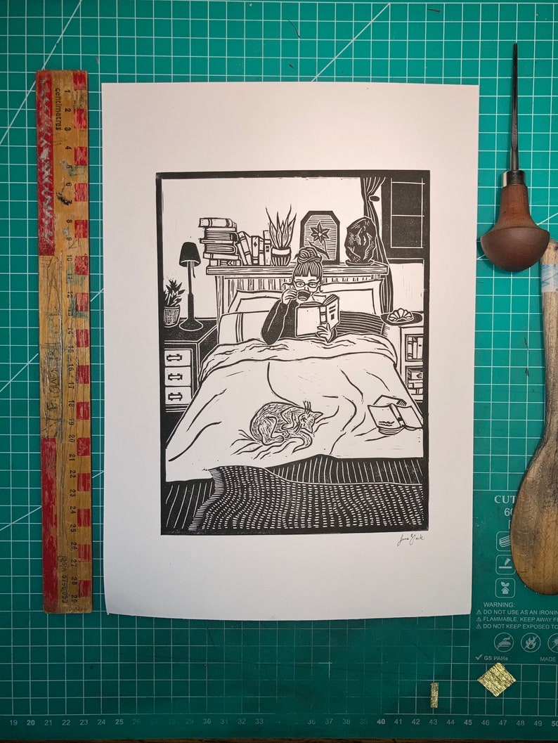 Peut inclure: Illustration en noir et blanc d'une personne lisant un livre au lit avec un chat enroul&eacute; sur la couverture. La personne porte des lunettes et tient une tasse de caf&eacute; ou de th&eacute;. Il y a des livres sur une &eacute;tag&egrave;re au-dessus du lit et une lampe sur une table de chevet.