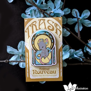 May include: A gold-colored enamel pin featuring a gray mouse with a floral crown and flowing blue ribbons. The pin is set against a yellow background with Art Nouveau-style lines and the word "TRASH" at the top. The card reads "Animal Nouveau" and "Enamel Pins by LABillustration."