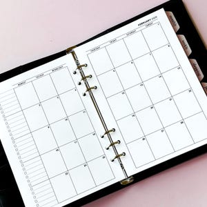 May include: A black ring-bound planner open to a February 2026 monthly calendar view. The white pages feature a grid layout with days of the week, numbered dates, and a vertical column of checkboxes on the left side. Black tabbed dividers are visible.