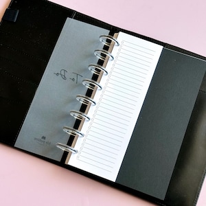 May include: Open black leather planner with a clear plastic overlay and lined paper. The overlay has the words "To Do" and a logo printed on it. The planner is open to reveal the lined paper and a black page.