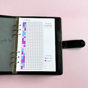 May include: A black leather ring-bound planner containing a Year in Pixels tracker page. The grid features columns for months and rows for days, with handwritten text Fitness Consistency and a color-coded legend for rest day, light walk, and workout.