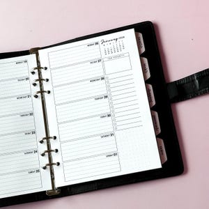 May include: A black ring-bound planner open to a weekly view for January 2026. The white pages feature black text, lined daily sections, a top priority box, and a checklist. Tabbed dividers labeled monthly, weekly, shopping, passwords, and notes.