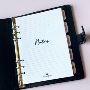 May include: A black leather planner with gold-colored rings, open to a lined page labeled "Notes." The planner has labeled tabs for "monthly," "weekly," "shopping," "passwords," and "notes." The Life Potion Planner logo is visible at the bottom.