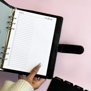 May include: A black six-ring binder planner featuring a white insert page with a checklist column and a dotted section labeled Brain Dump. The binder has a metal ring mechanism and a side strap closure, with black tabbed dividers visible nearby.